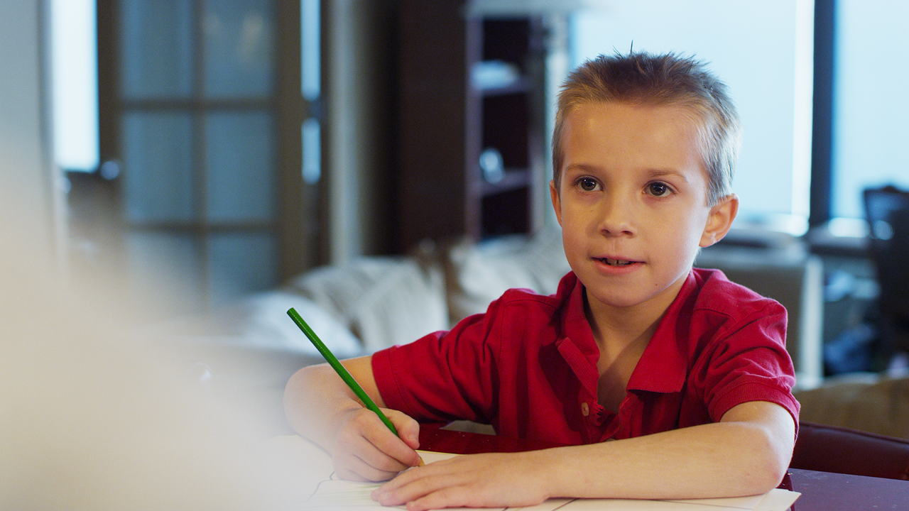 Young boy draws in a notebook while talking to someone offscreen.