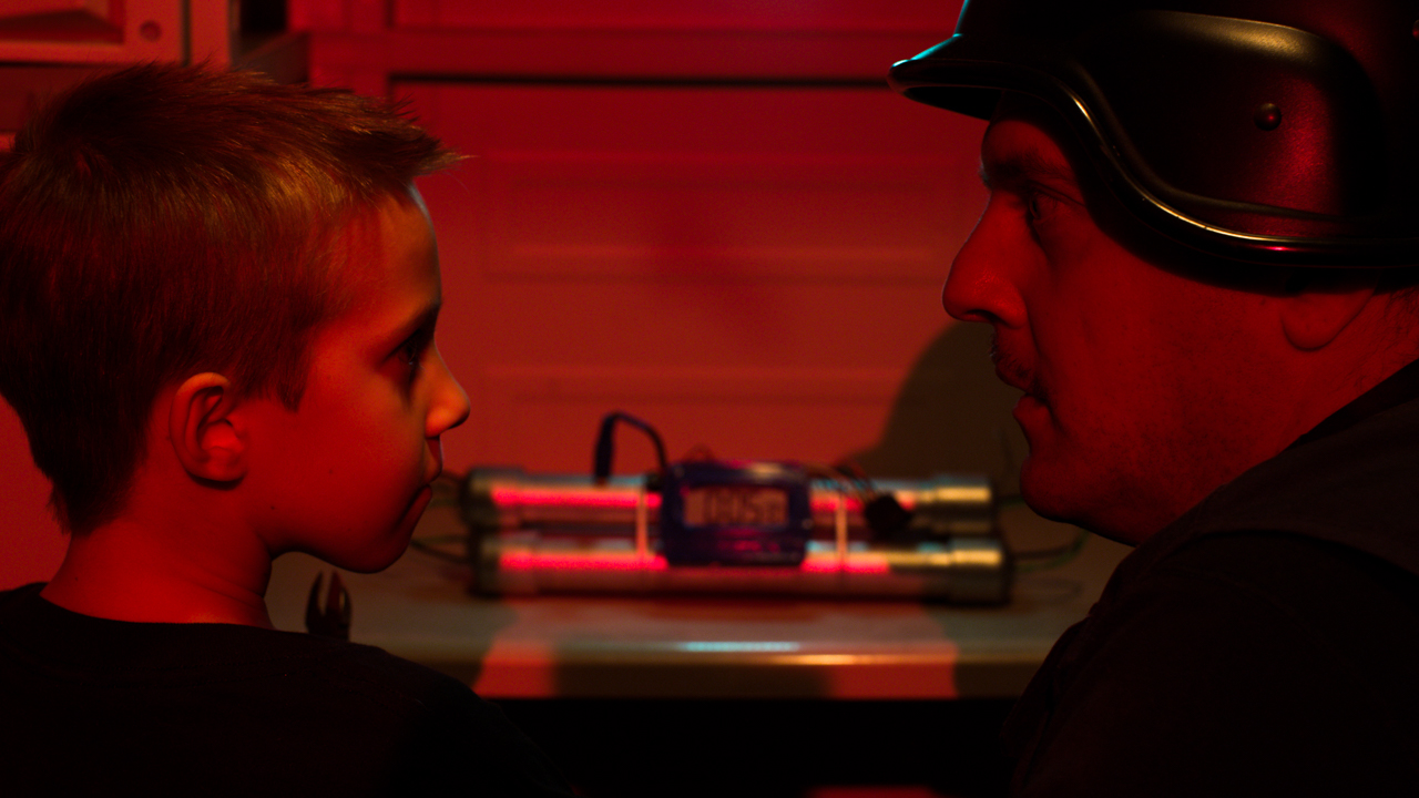 Father and son talking in a red lit room with bomb squad outfit and explosive device.