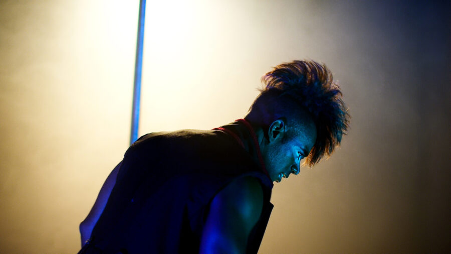 Man with mohawk hairstyle pole dances onstage with yellow light in clown makeup.