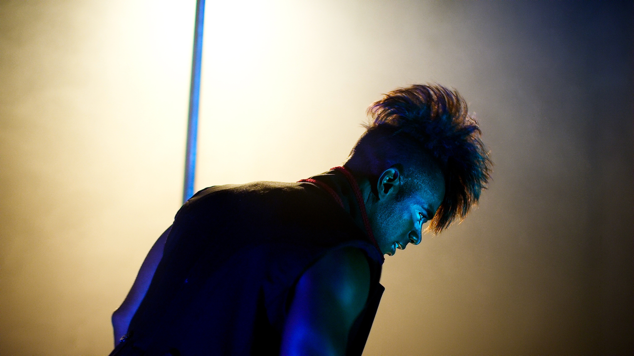 Man with mohawk hairstyle pole dances onstage with yellow light in clown makeup.