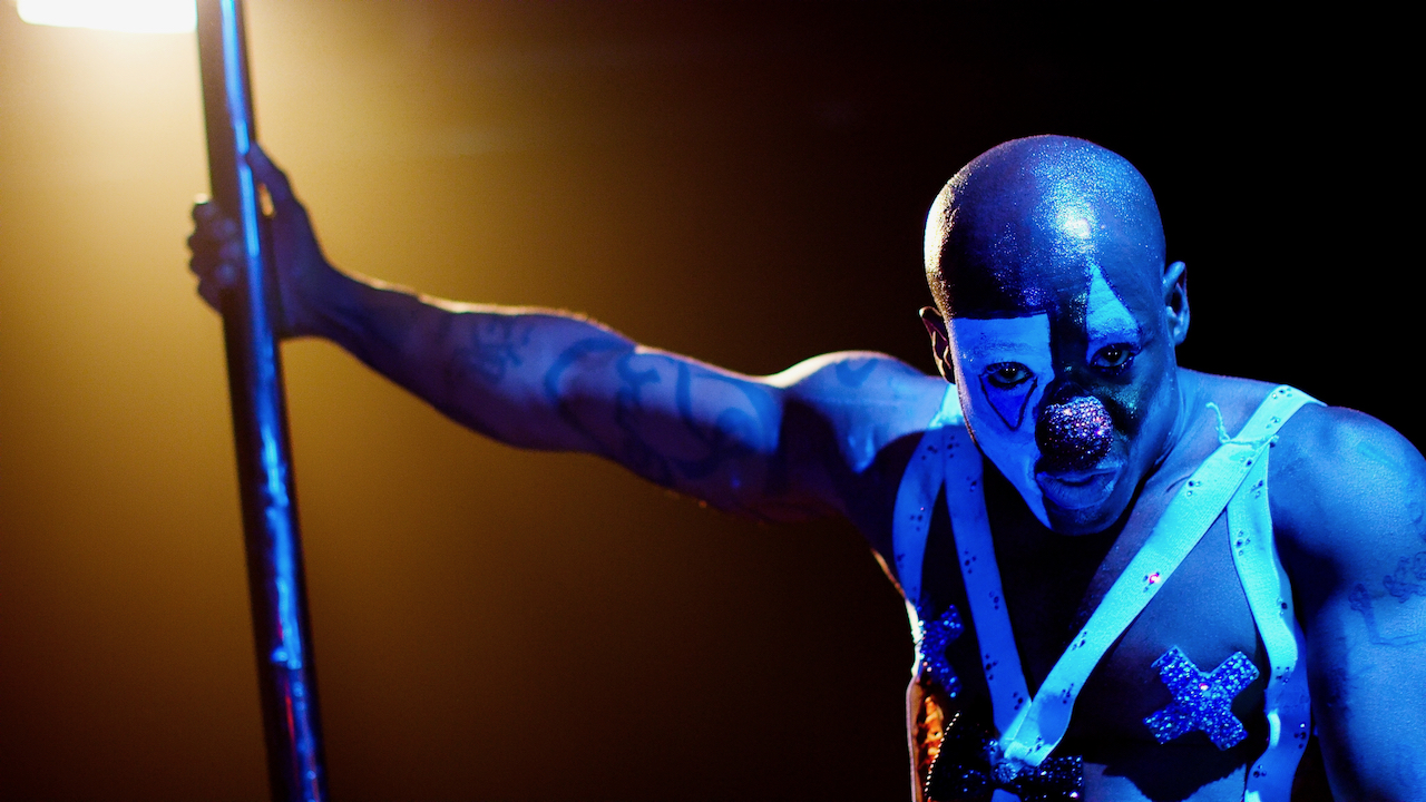 Man pole dances onstage with yellow and blue light in clown makeup.