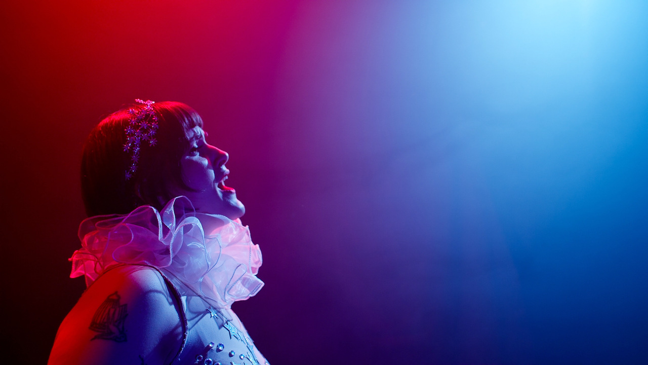 Woman with short dark hair looks sings in blue and red light in a clown costume.