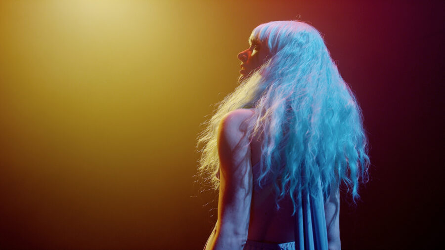 Woman with long hair looks behind in yellow, and orange light and clown makeup.