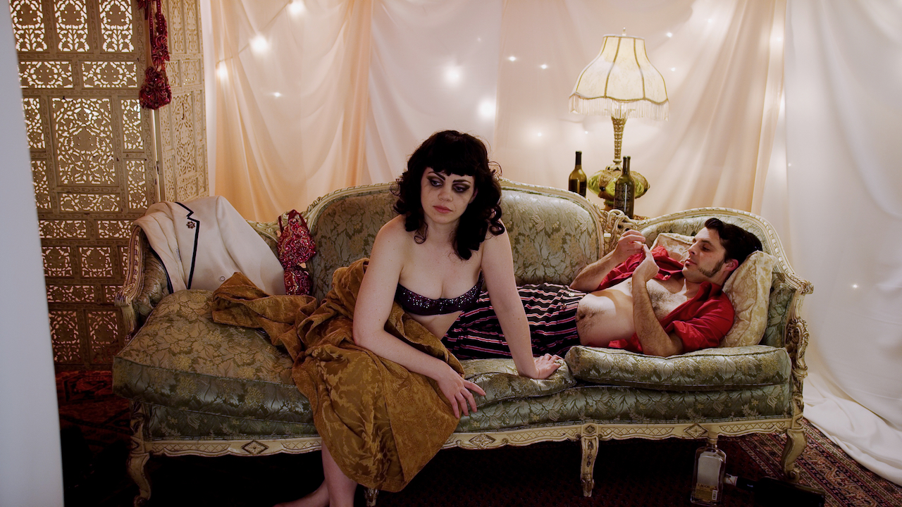 Woman with long black hair sits on fancy couch looking sad in a white tented room with ball lights.