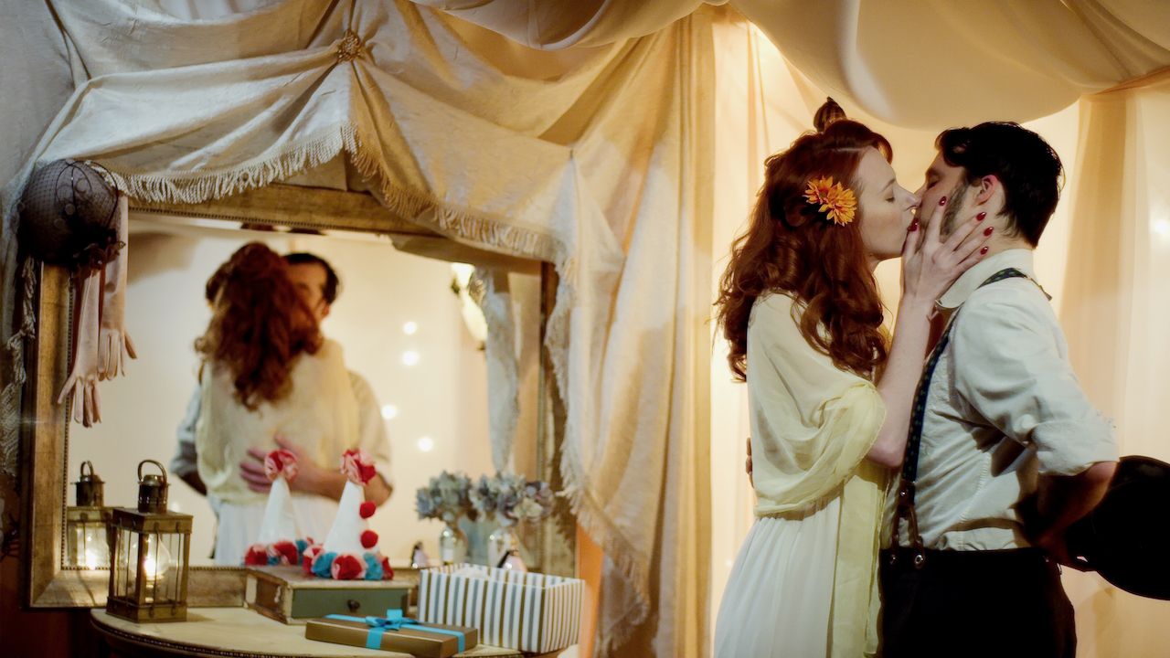 Woman with long red hair kisses man with sideburns in a yellow tented room with mirror.