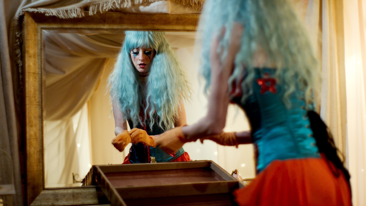 Woman with long blue hair and clown makeup takes off gloves in a yellow tented room.