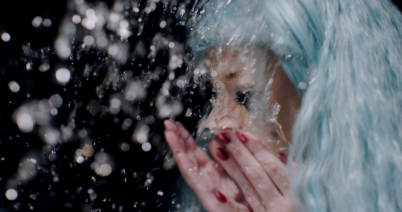 Woman in long blue hair splashes water on her face in slow motion.
