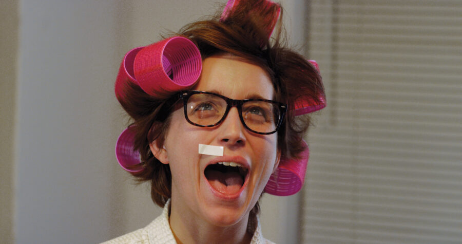 Woman in pink hair curlers and turtle shell glasses screams with a beauty strip on her face wearing a white robe.