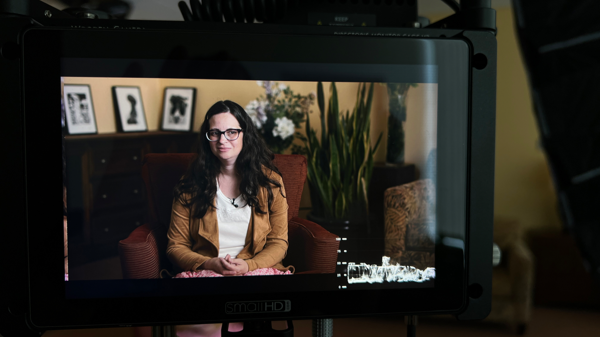 Monitor shot of a woman with glasses being interviewed in a yellow colored room with large green plants.