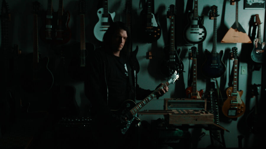 man in shadows plays guitar in front of wall of hung guitars