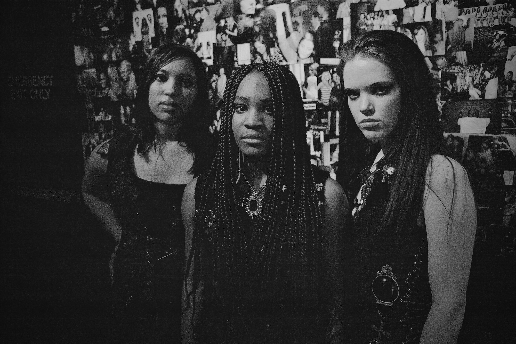 Girls stand backstage in rockstar outfits with a postered wall in black and white color.