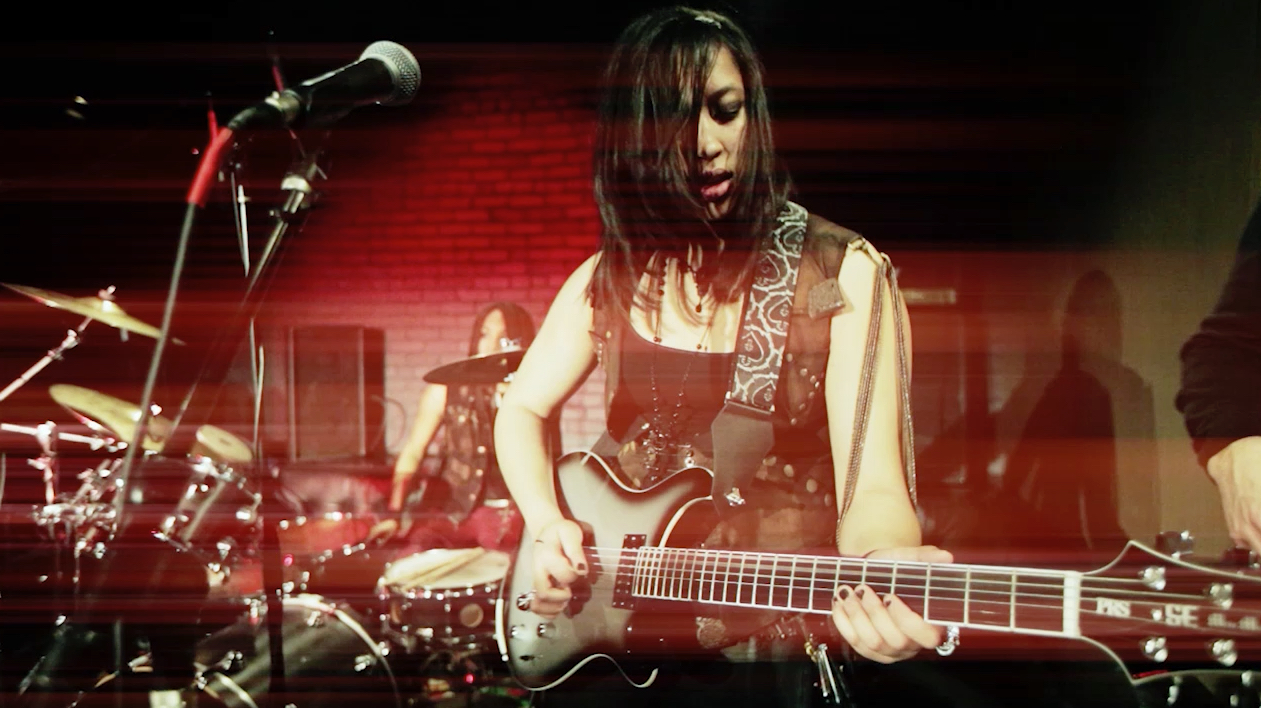 Girl with long black hair and guitar onstage performing in red light.