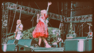 Female singer with long white and pink punk dress raising hand on microphone.