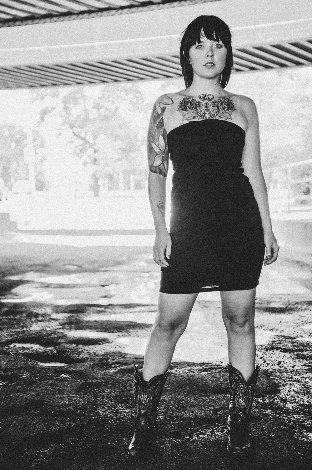 Black and white photo of girl in cowboy boots, tattoos, and a black dress under a bridge.