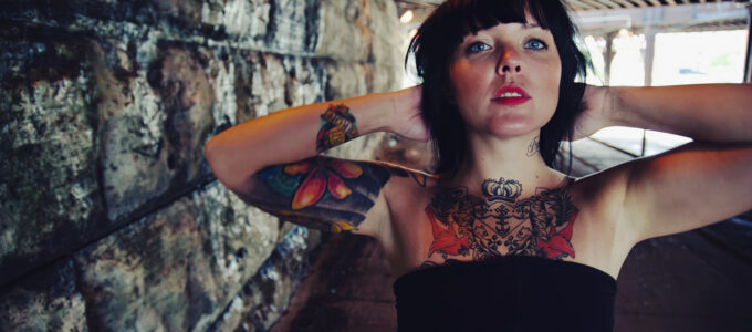 Photo of girl with black hair, tattoos, and black dress under a bridge.