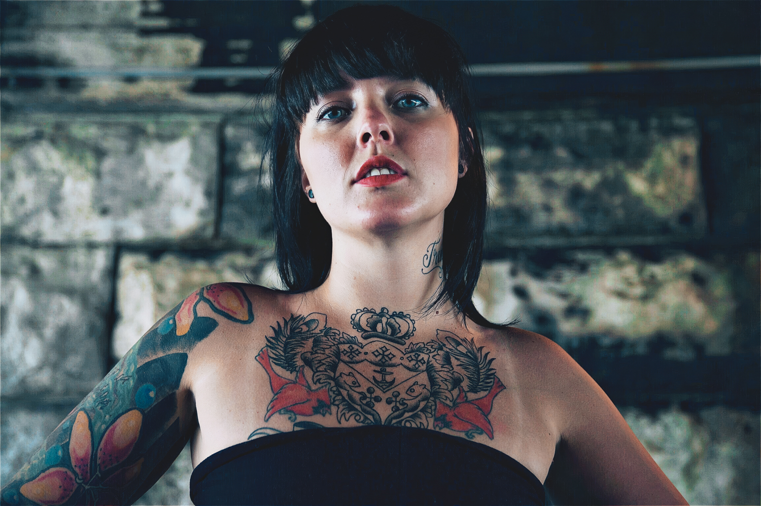 Photo of girl with black hair, tattoos, and black dress under a bridge.