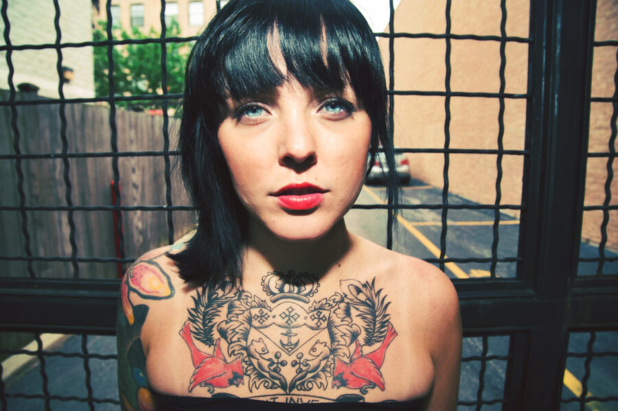 Photo of girl with black hair, tattoos, and black dress in front of a gated fence.