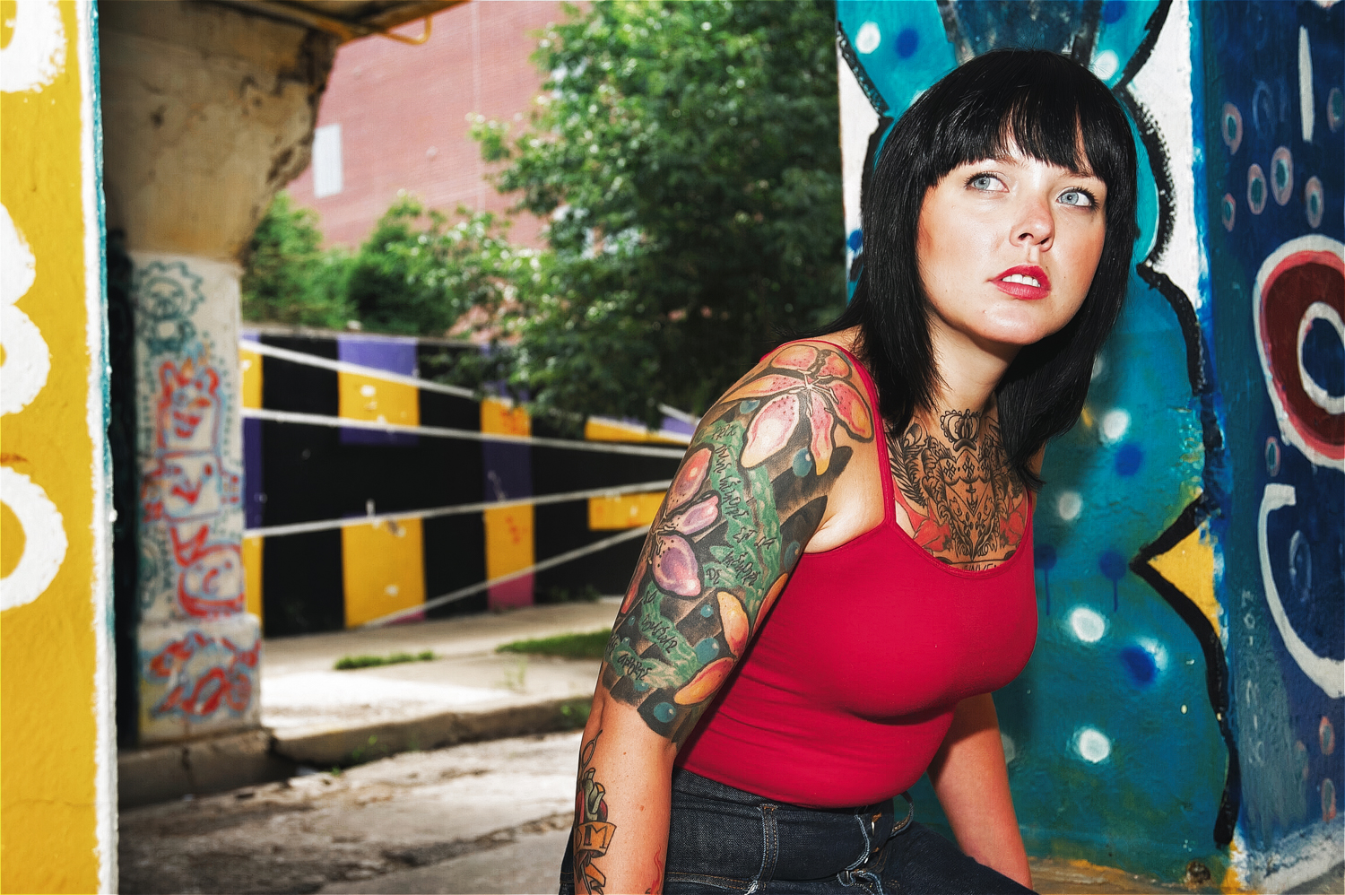 Photo of girl with black hair, tattoos, and red top under a painted mural bridge.