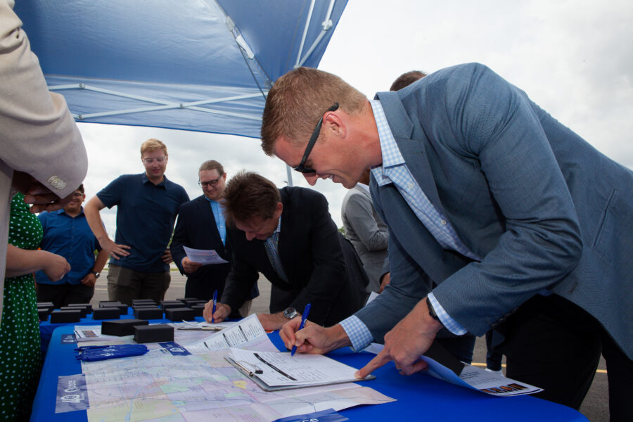 Men sign a paper register at a table under a umbrella.