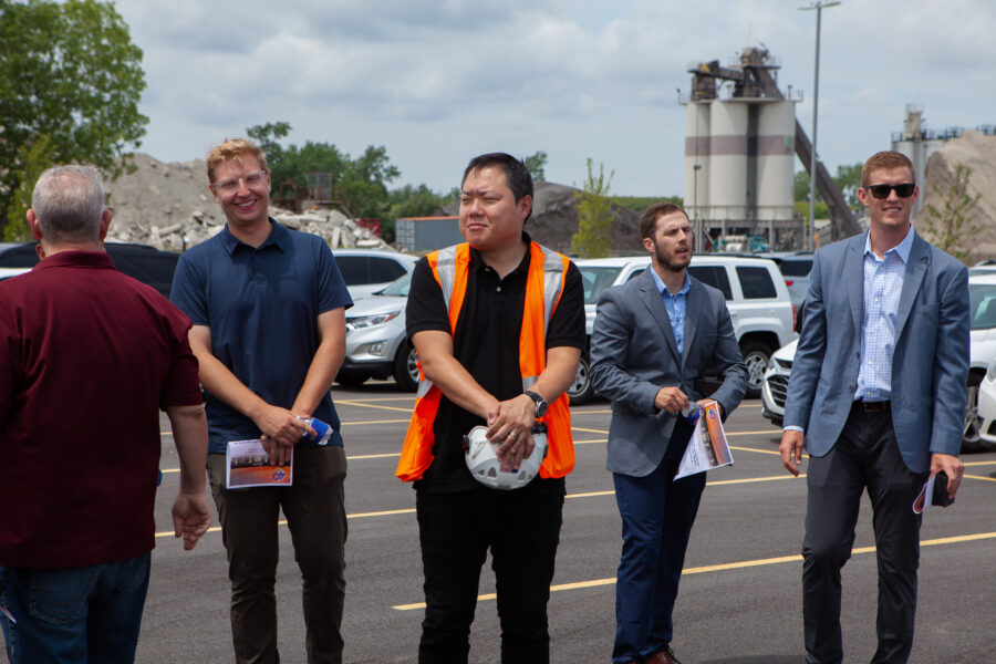 Construction workers stand in a parking lot with a factory in the distance.