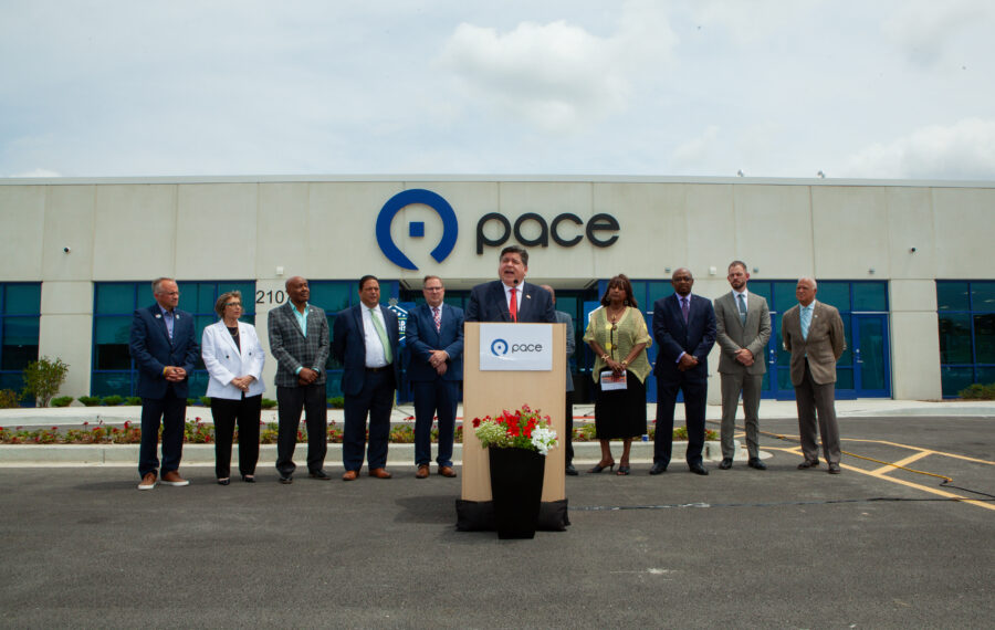 Man at podium gives speech in a parking lot surrounded by people in suits.