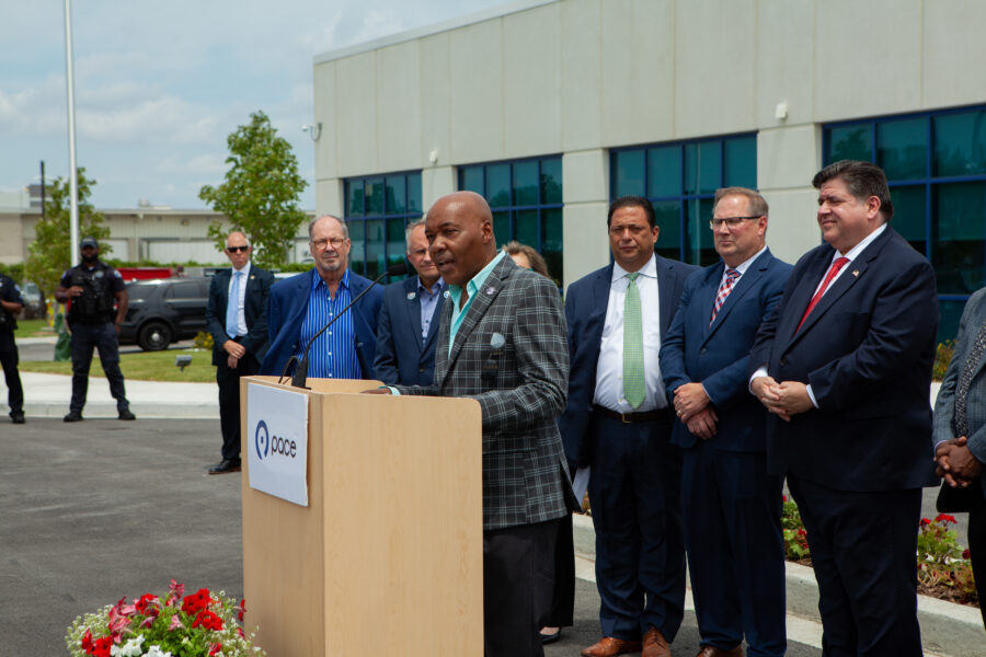 Man at podium gives speech in a parking lot surrounded by people in suits.