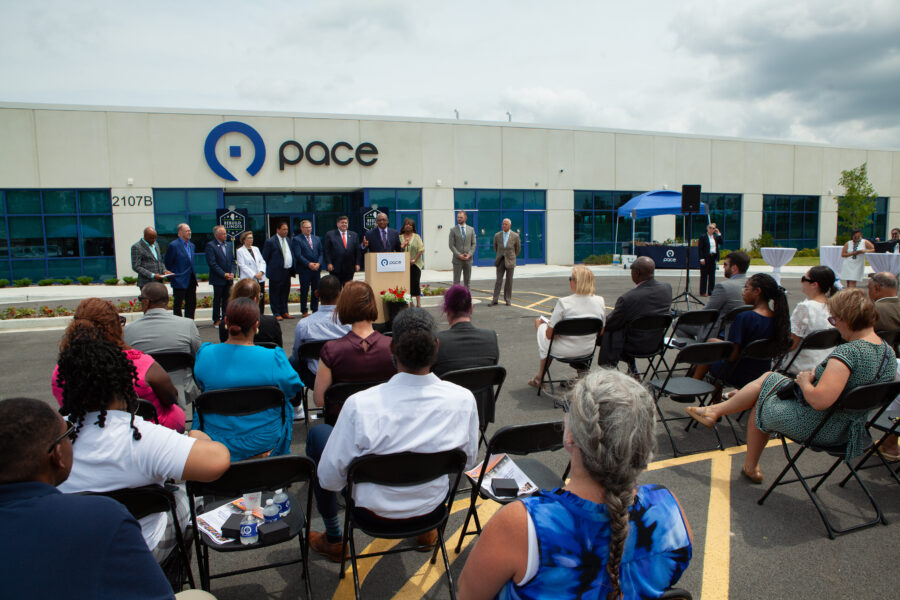 Man at podium gives speech in a parking lot surrounded by people in suits and a large crowd sitting.