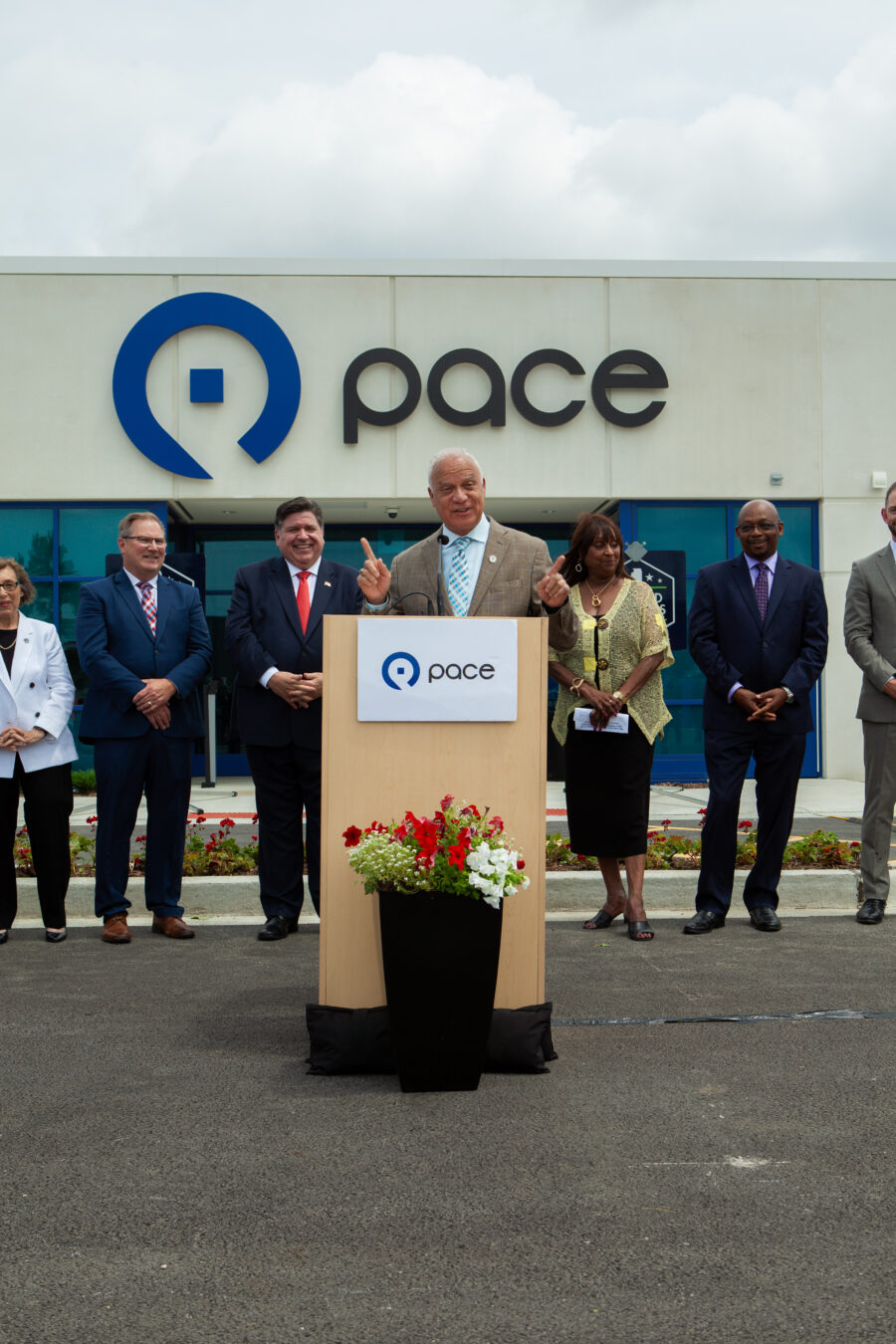 Man at podium gives speech in a parking lot surrounded by people in suits.