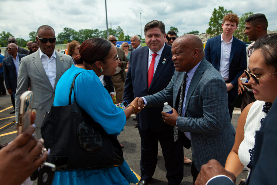 Smiling man shakes hands with woman in a blue dress in a parking lot surrounded by people.