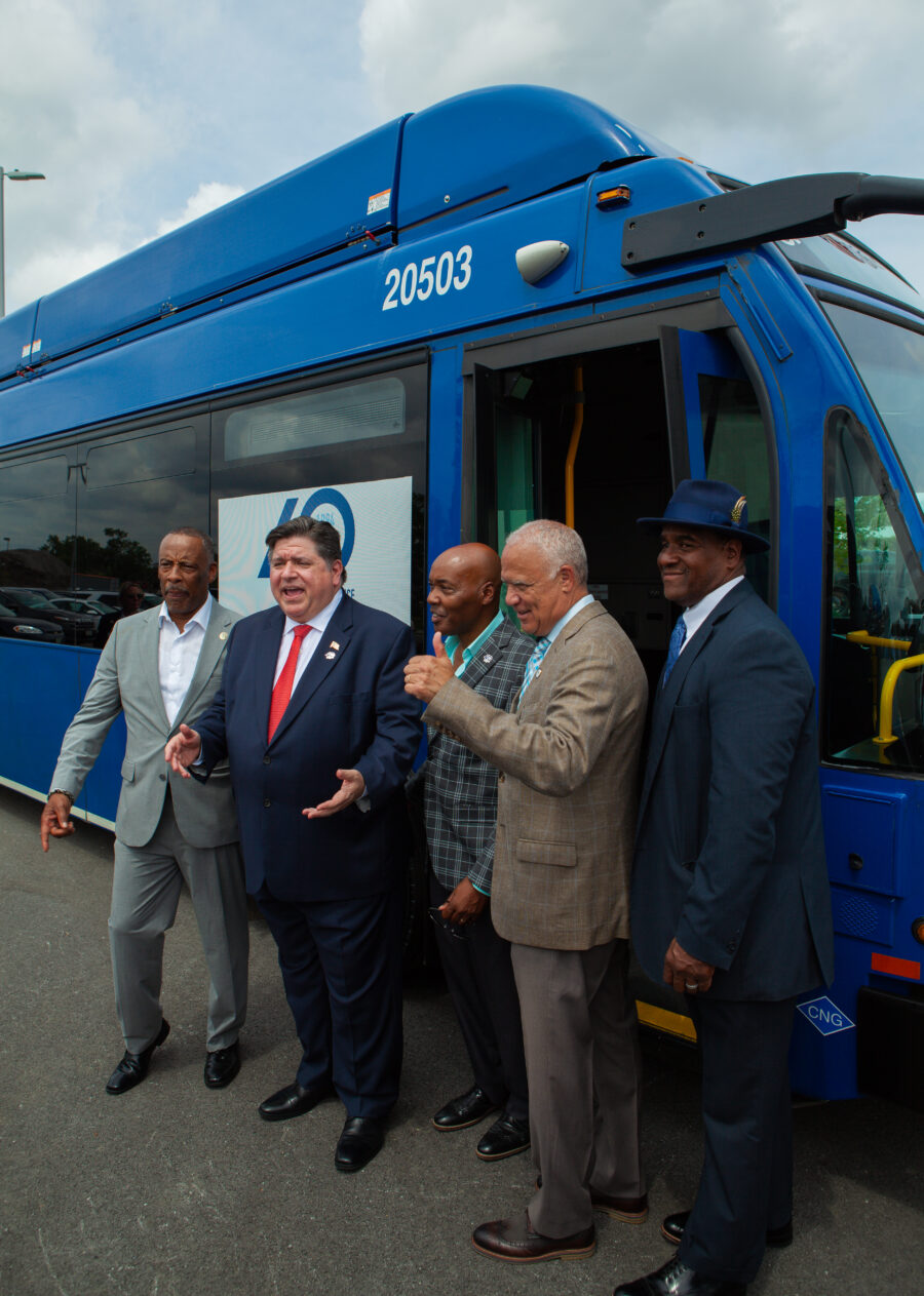 Men smiling and standing next to a bus give funny gestures off camera.