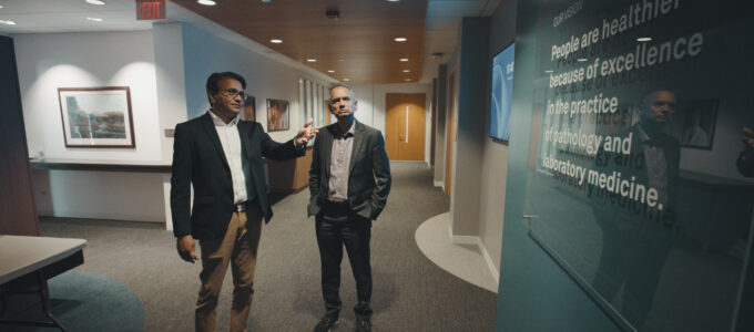 Two men talk with each other looking at green sign in suits.