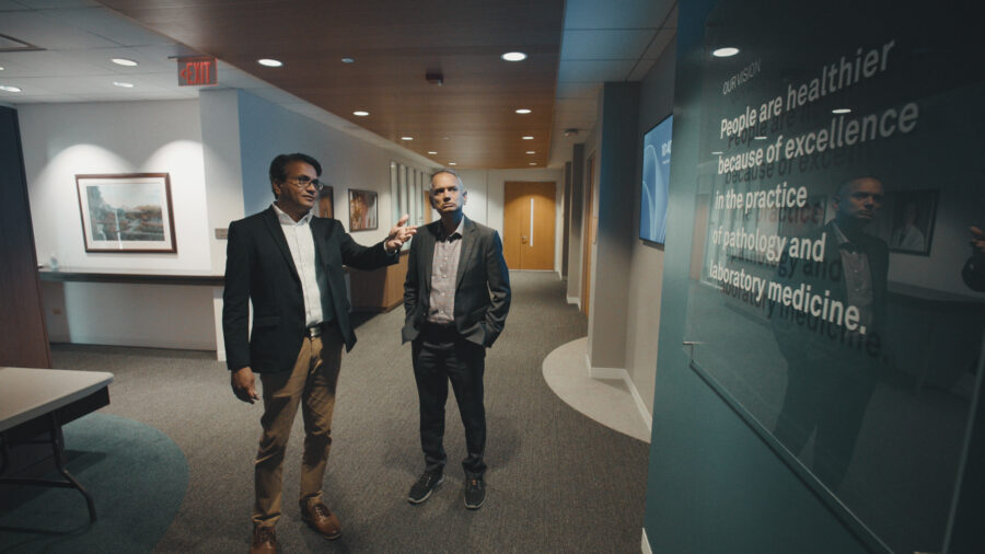 Two men talk with each other looking at green sign in suits.