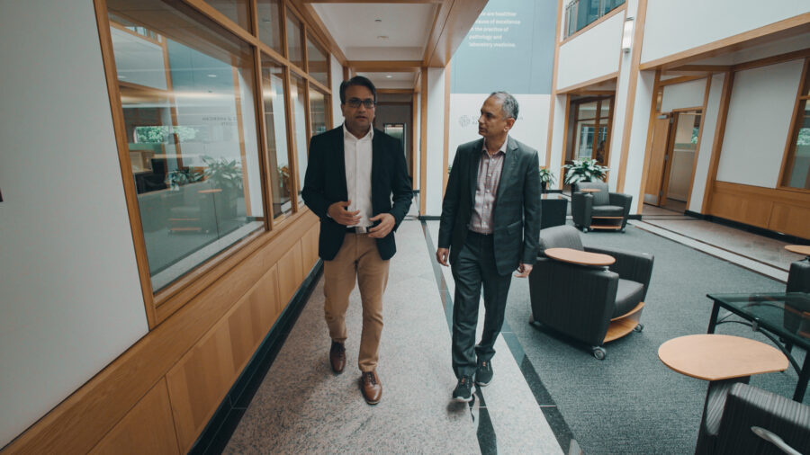 Two men walk and talk with each other in a large open lobby in suits.