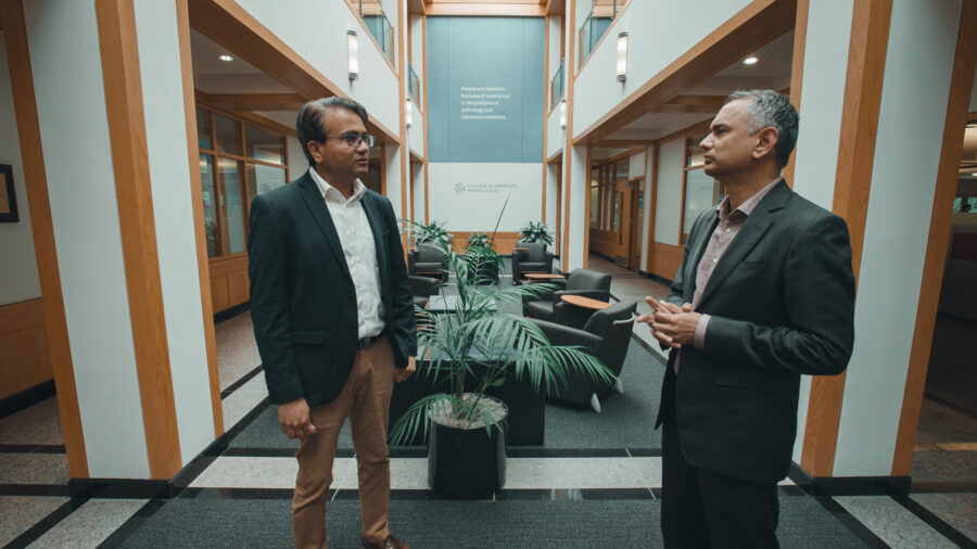 Two men talk with each other in a large open lobby in suits.