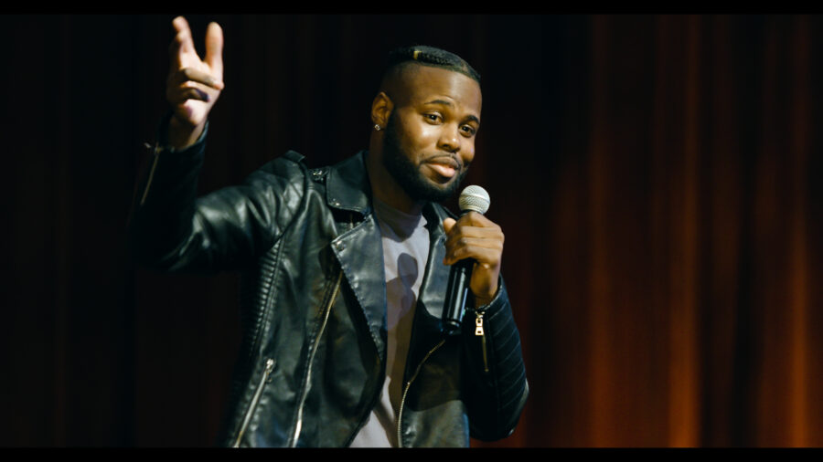 stand up comedy performer onstage with microphone and hand raised