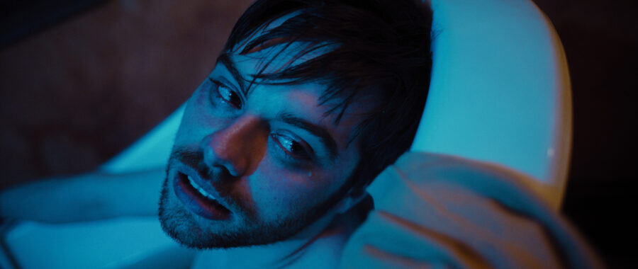 Man in blue light lays inside bathtub looking over towards the camera.