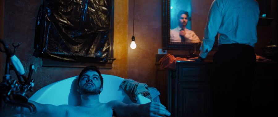 Man sits in tub full of water and blue light looking tired as man in background looks into mirror.