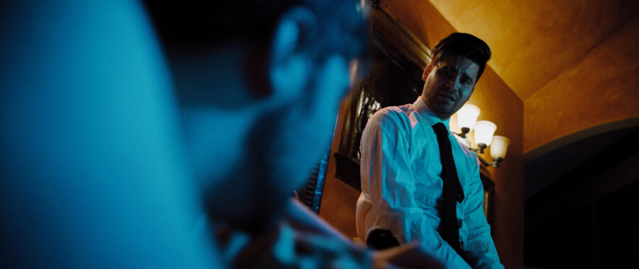 Man sits on bathroom sink looking towards another man in a bathtub with blue and yellow lights.