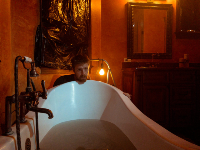 Man with face on a bathtub in a dimly lit room with garbage bags over the windows.