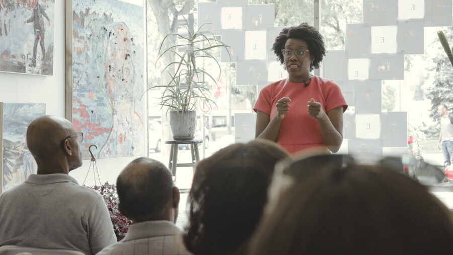 Woman in pink dress standing giving a speech to a crowd of people in a art gallery.