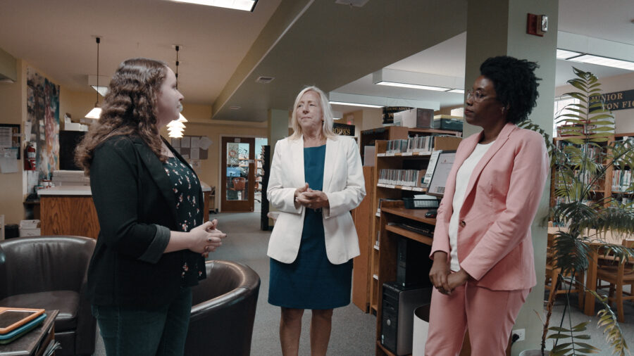 Woman in pink suit talks to 2 other women inside a library.