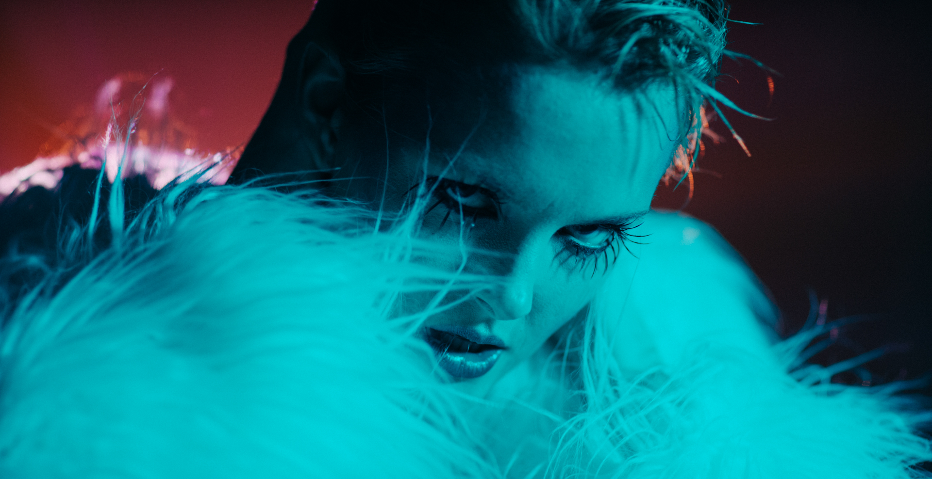 model in teal light stands in front of a red background in a white fur coat