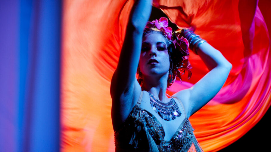 Gypsy woman twils orange silks in blue light with red flowers.