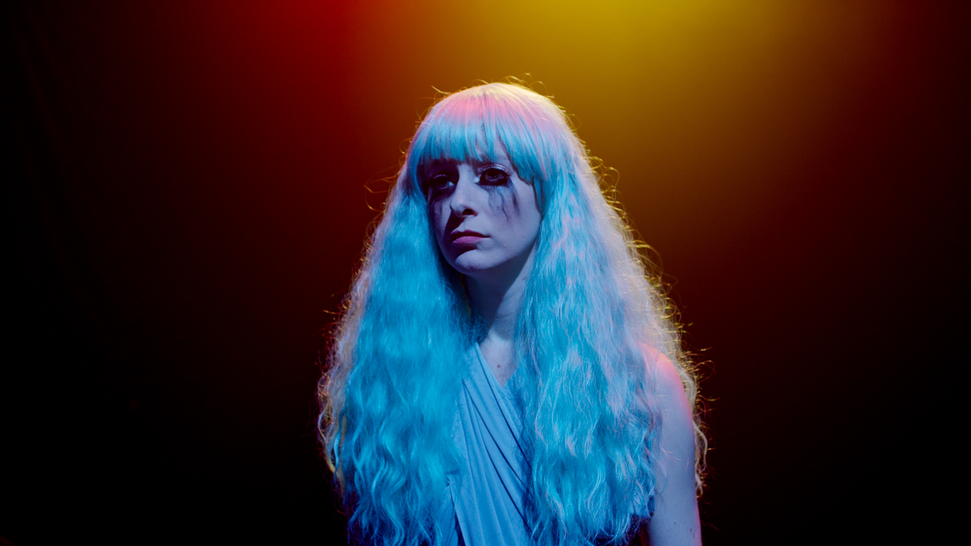 Woman with long hair looks sad in blue, yellow, and orange light and clown makeup.