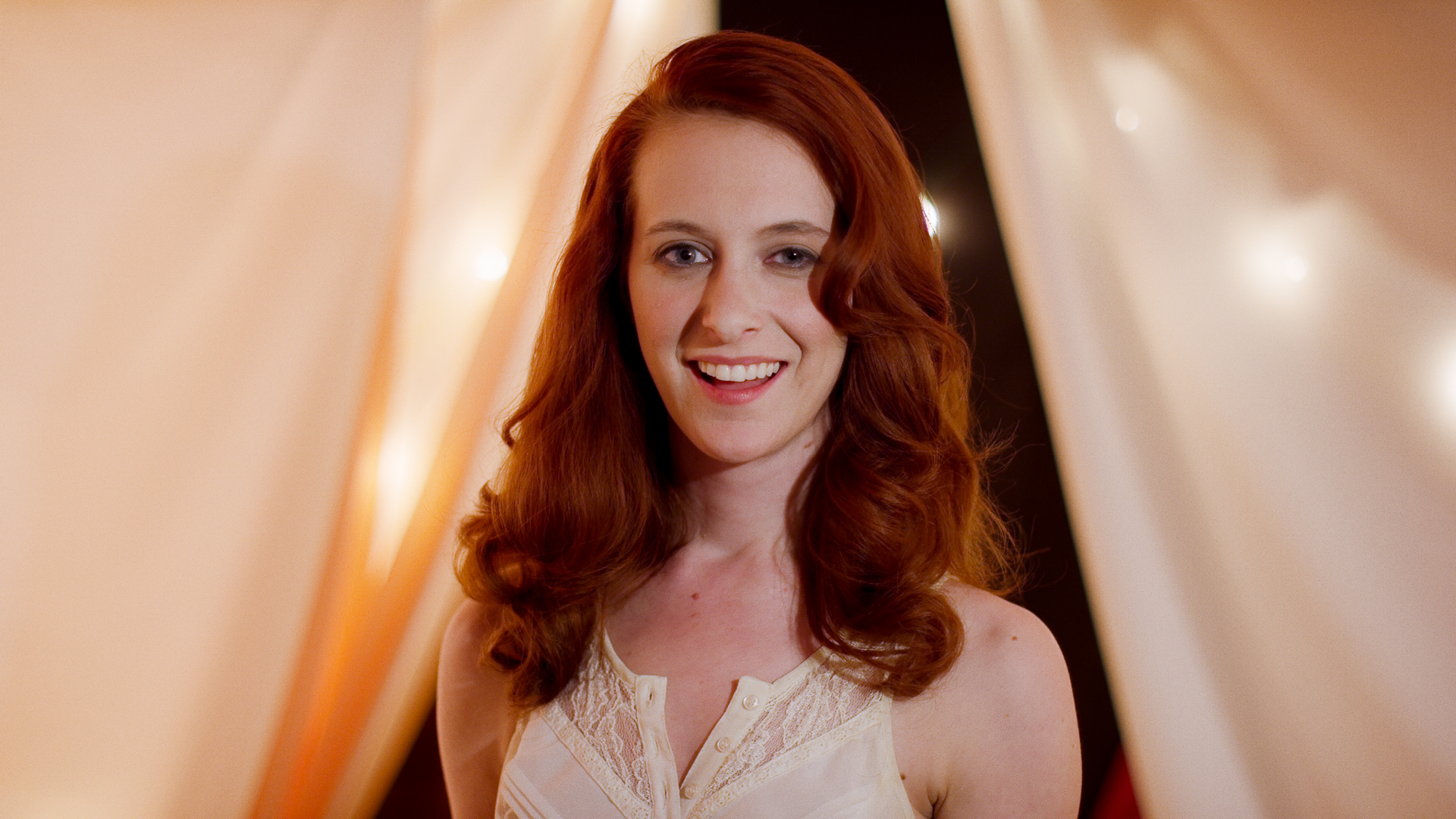 Woman with long red hair smiles at camera in a white tented room with ball lights.