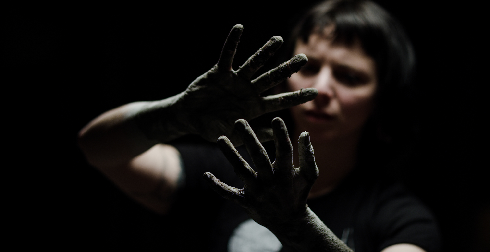 Woman in spotlight looks at hands covered in mud.
