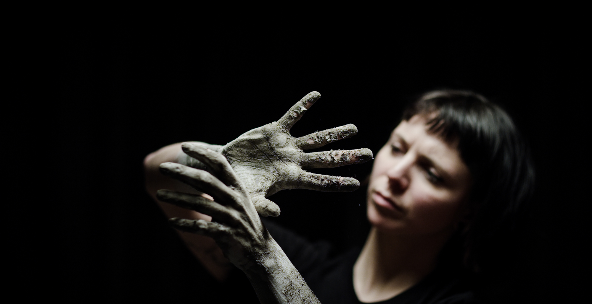 Woman in spotlight looks at hands covered in mud.