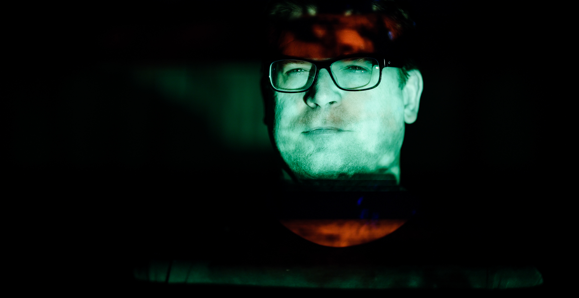 Man in glasses stands with projector video shining on face.