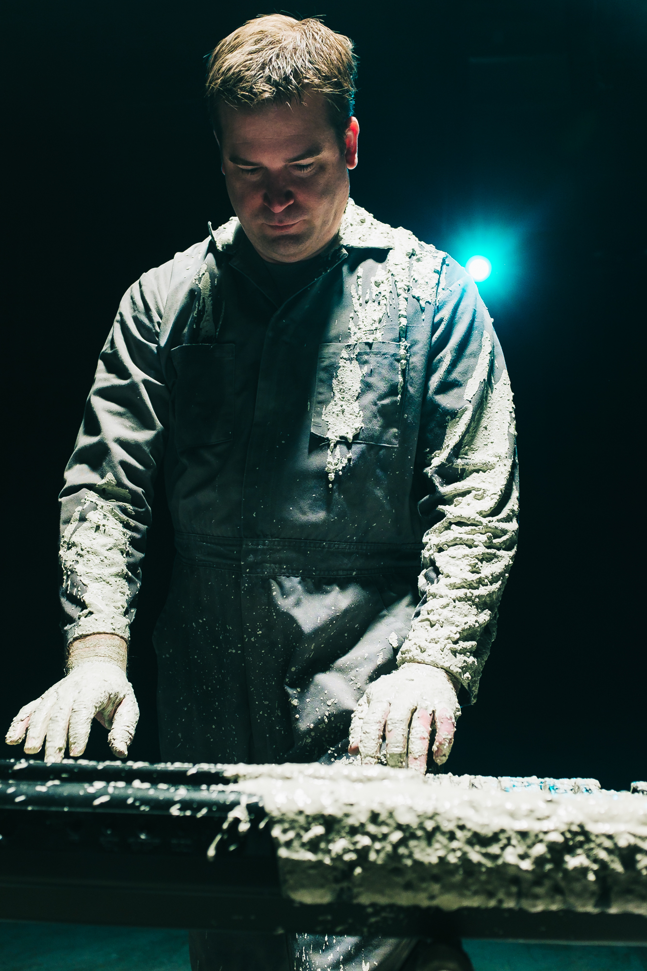 Man covered in mud dressed in overalls plays keyboard lit from above in black room.
