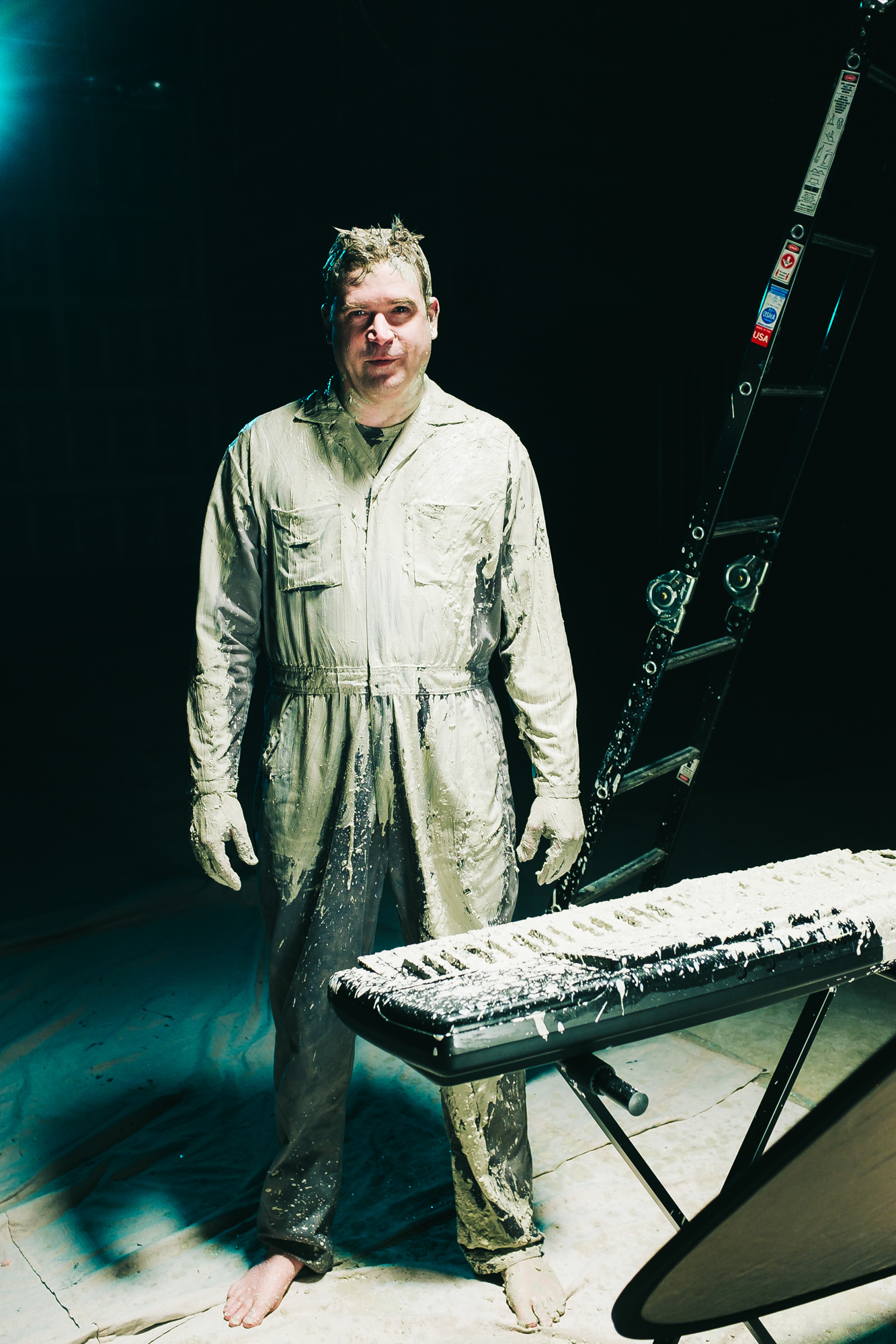 Man covered in mud dressed in overalls next to a keyboard and ladder.